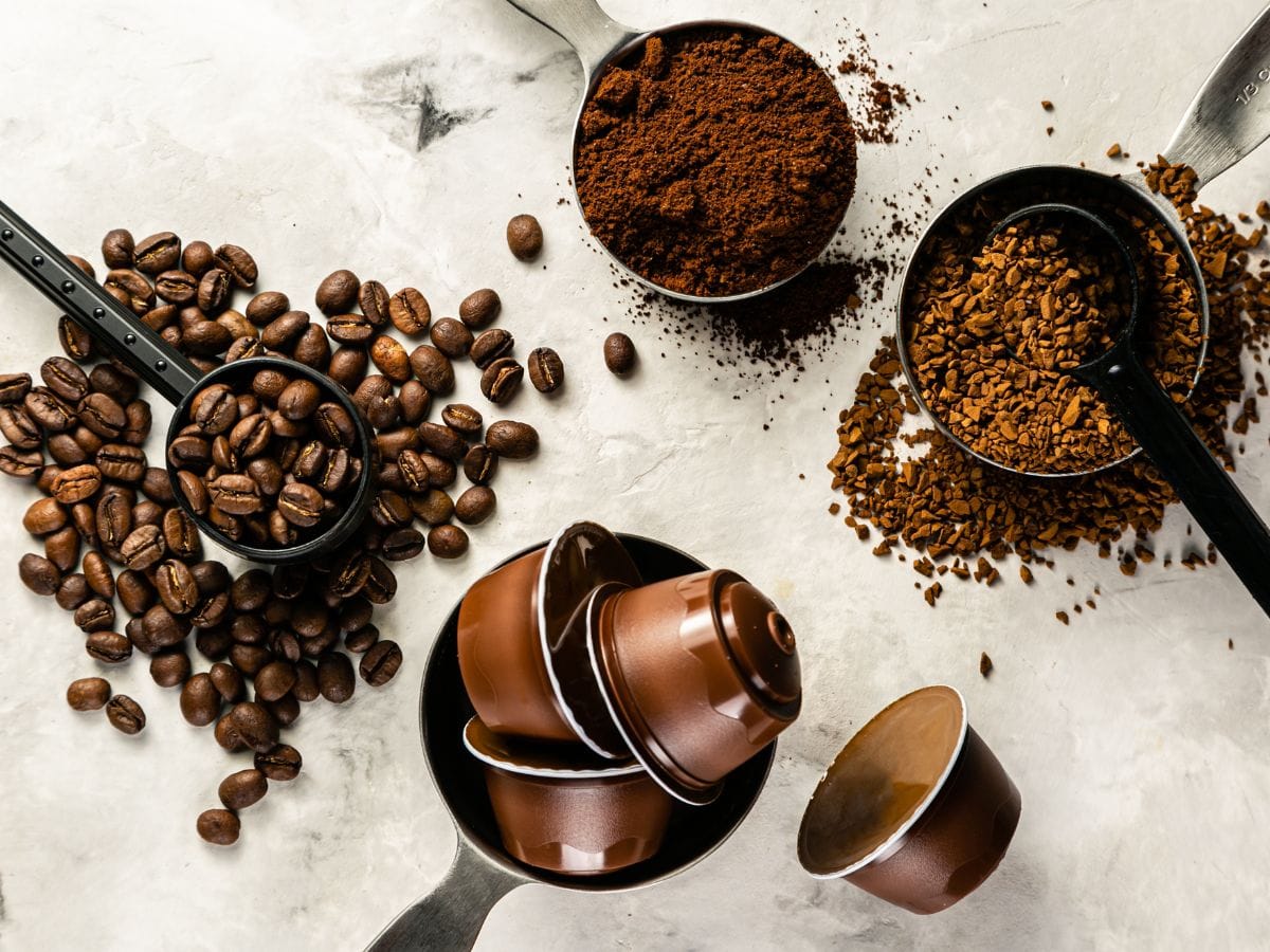Variedad de formatos de café de especialidad con granos de café enteros, café de especialidad molido en cuchara medidora y cápsulas sobre fondo claro.