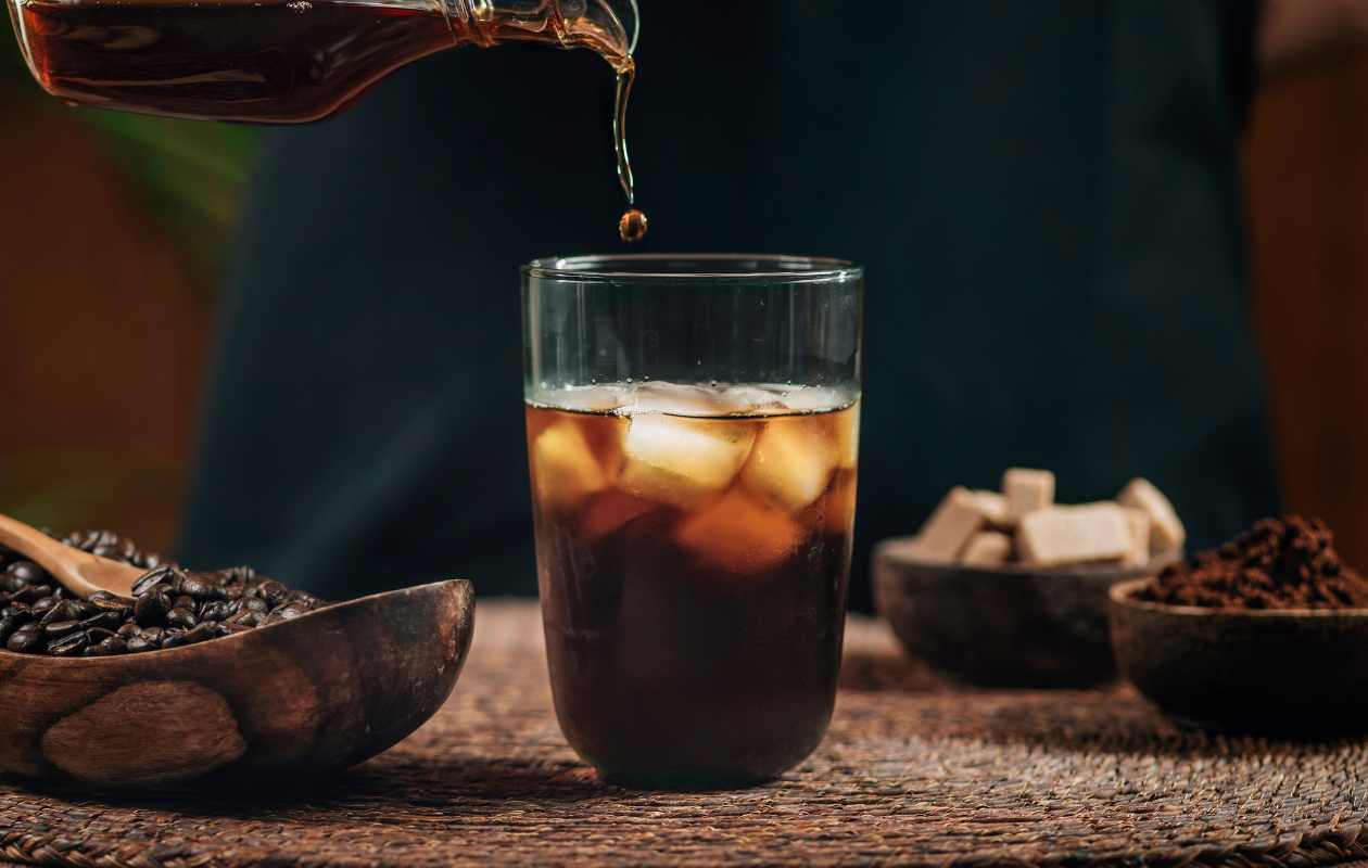 Vaso con café Cold Brew y cubitos de hielo sobre una mesa de madera rústica, con granos de café al fondo - Memento Coffee